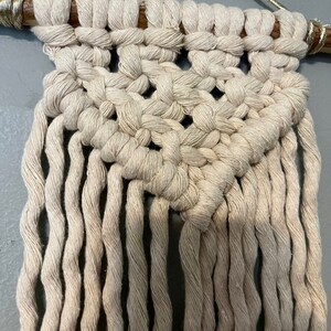 Op de afbeelding: Een beige macramé wandhanger met een geometrisch patroon en franjes. De hanger is gemaakt van natuurlijke katoenen koord en is bevestigd aan een houten stok.