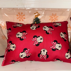 May include: A red rectangular cushion adorned with a pattern of Minnie Mouse faces, each with a red and white polka dot bow. This cushion is a decorative item, suitable for a child's room or festive occasions.