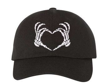 May include: Black baseball cap featuring a white embroidered design of two skeleton hands forming a heart shape. The cap has a curved brim and a structured crown. This hat is a unique accessory for those who love gothic or alternative fashion.