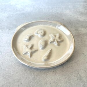 May include: A cream-colored ceramic dish with embossed seashell designs. The dish features a variety of sea creatures, including starfish, seashells, and a conch shell. The dish is round with a slightly raised edge.