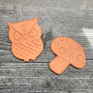 May include: Two terracotta-colored ceramic figurines: an owl and a mushroom. The owl has large eyes and a textured body. The mushroom has a rounded cap with circular indentations. Both are likely garden decor.