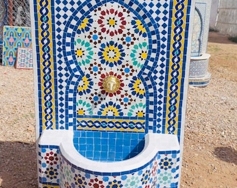May include: A white ceramic wall fountain with a blue tiled basin and a decorative archway. The archway is decorated with intricate geometric patterns in shades of blue, yellow, red, and green. A gold faucet is visible at the bottom of the archway.