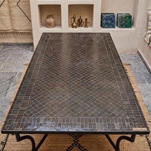 Peut inclure: Une table rectangulaire dotée d'un plateau en mosaïque gris foncé avec un motif en losange au centre et une bordure de carreaux rectangulaires. La table est soutenue par un cadre en métal noir aux pieds incurvés et porte une petite étiquette.
