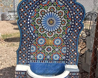 May include: A decorative wall fountain with a blue, white, red, and green mosaic tile design. The fountain has a curved basin and a spout in the center.