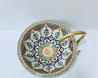 May include: A decorative ceramic bowl with a detailed floral pattern in white, blue, yellow, and green. It features a brass handle and base, and a light green rim. The bowl is likely for decorative purposes.