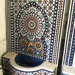 May include: A white tiled fountain with a blue and yellow mosaic design. The fountain has a curved basin and a spout with a gold finish.