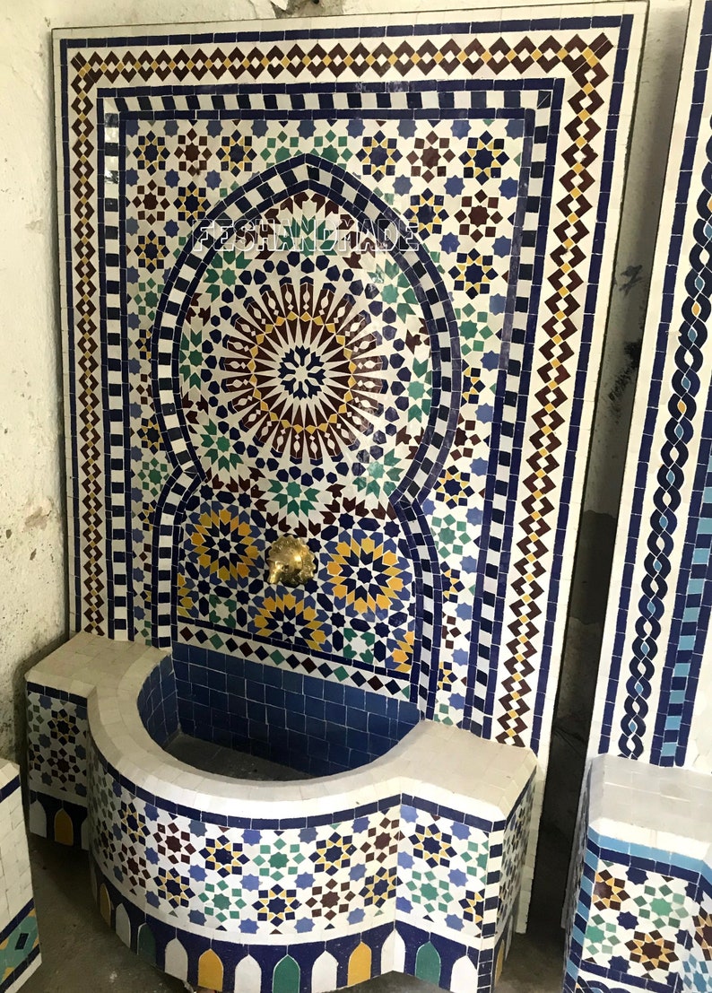 May include: A white tiled fountain with a blue and yellow mosaic design. The fountain has a curved basin and a spout with a gold finish.