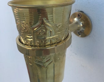May include: A gold-colored metal toothbrush holder with an ornate design. The holder is mounted to a wall.