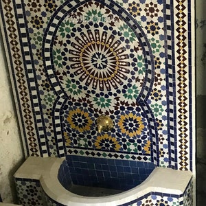 May include: A white ceramic fountain with a blue and white mosaic design. The fountain has a curved basin and a spout in the center. The mosaic design features geometric patterns and stars.