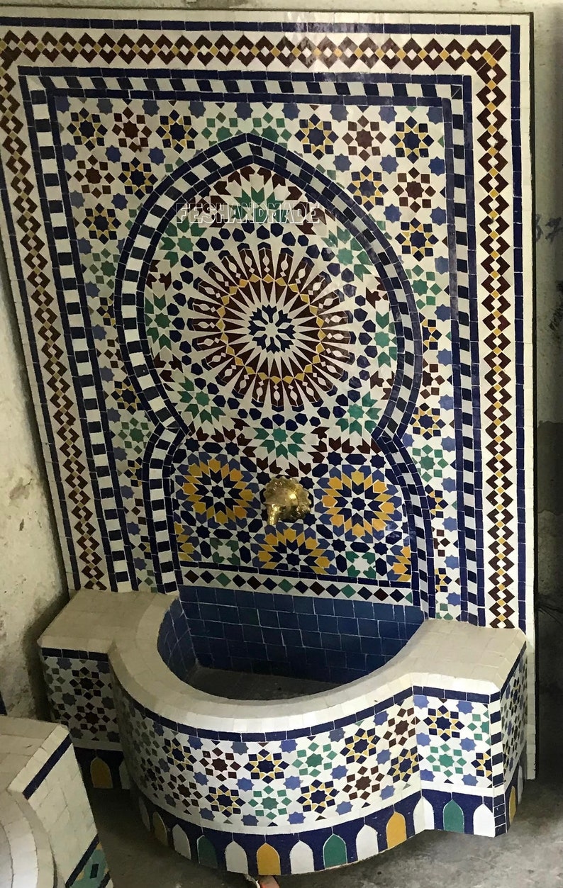 May include: A white ceramic fountain with a blue and white mosaic design. The fountain has a curved basin and a spout in the center. The mosaic design features geometric patterns and stars.