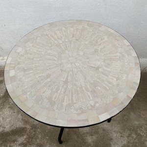 Puede incluir: Una mesa redonda de mosaico blanco con un patrón geométrico. La mesa tiene una base de metal negro con tres patas.
