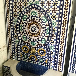May include: A white ceramic fountain with a blue and white mosaic design. The fountain has a traditional Moroccan arch and a spout with a gold finish. The fountain is set against a white wall.