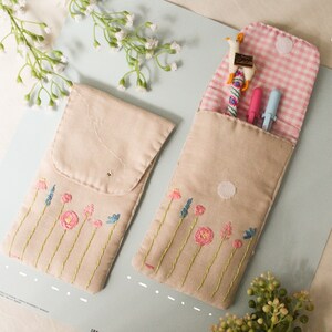 May include: Two beige fabric pencil cases with embroidered flowers. One case is open and has a pink and white checkered lining. The case contains a blue and pink pen, a blue pen, and a small toy llama.