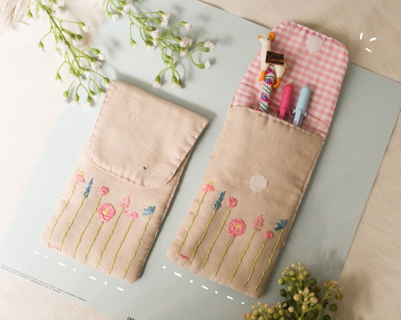 May include: Two beige fabric pencil cases with embroidered flowers. One case is open and has a pink and white checkered lining. The case contains a blue and pink pen, a blue pen, and a small toy llama.