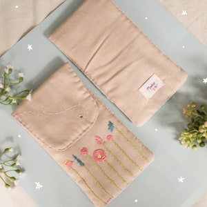 May include: Two beige fabric pouches with embroidered flowers and a bee on one pouch. The pouch on the left has a flap with a stitched closure. The pouch on the right has a label that says "Made with Love".
