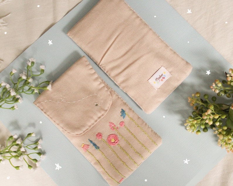 May include: Two beige fabric pouches with embroidered flowers and a bee on one pouch. The pouch on the left has a flap with a stitched closure. The pouch on the right has a label that says "Made with Love".