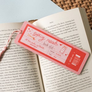 May include: A pink and red bookmark with the text "Spicy Reads Book Club" and a drawing of a lit matchstick. The bookmark also has a ticket design with the text "Admit One" and a barcode.