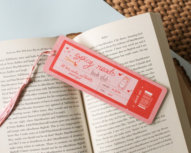 May include: A pink and red bookmark with the text "Spicy Reads Book Club" and a drawing of a lit matchstick. The bookmark also has a ticket design with the text "Admit One" and a barcode.