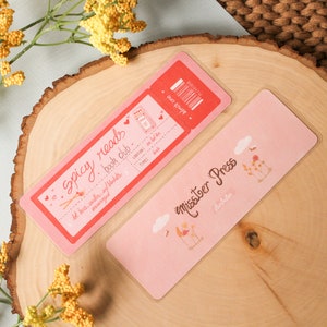 May include: Two pink bookmarks. One bookmark is a ticket stub with the text "Spicy Reads Book Club" and the other bookmark has a floral design and the text "Miss.toer Press".