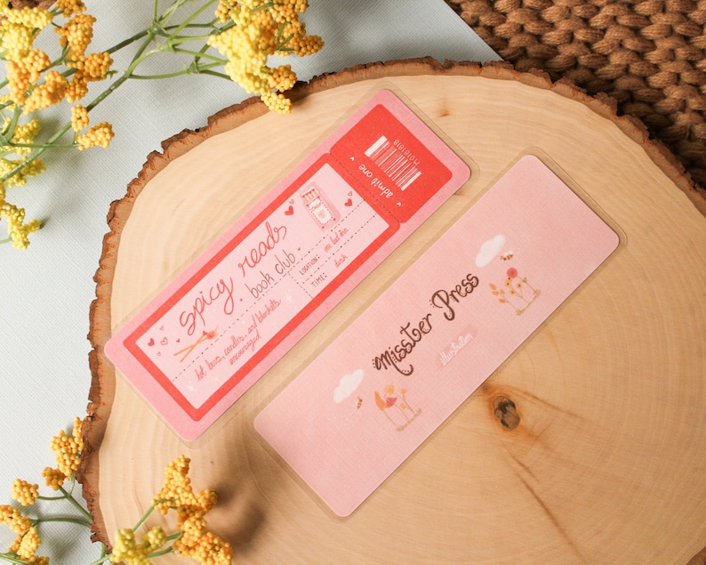 May include: Two pink bookmarks. One bookmark is a ticket stub with the text "Spicy Reads Book Club" and the other bookmark has a floral design and the text "Miss.toer Press".