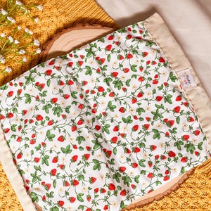 May include: A white fabric bag with a strawberry print and a beige fabric border. The bag has a small label with the text "Made with Love" on it.