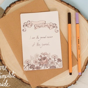 May include: A blank journal with a floral design and the text "Solemnly swear I am the proud owner of this journal." on a brown envelope with two pens on a wooden surface.