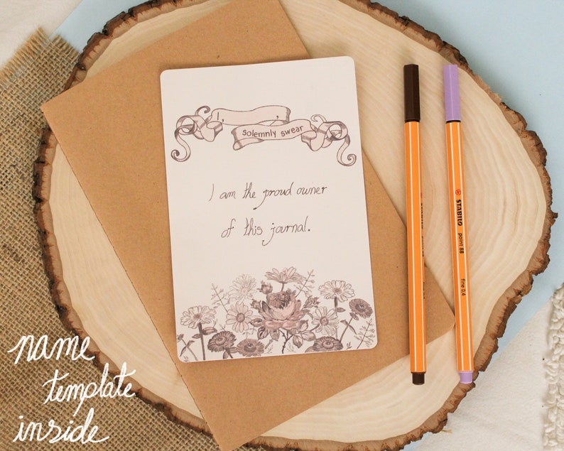 May include: A blank journal with a floral design and the text "Solemnly swear I am the proud owner of this journal." on a brown envelope with two pens on a wooden surface.