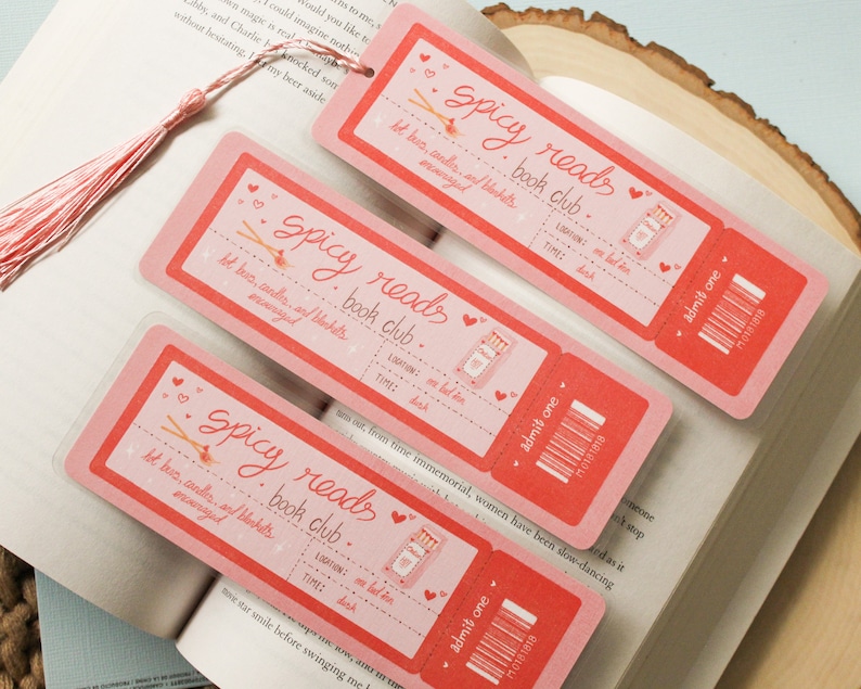 May include: Three pink bookmarks with a whimsical design featuring a book, matches, and the text "Spicy Reads Book Club". The bookmarks have a ticket-style design with the text "Admit One" and a barcode.