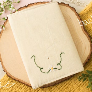 May include: A white canvas book cover with a green embroidered floral design. The cover is sitting on a wooden slice.