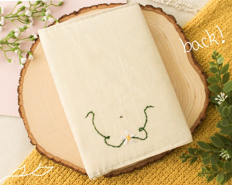 May include: A white canvas book cover with a green embroidered floral design. The cover is sitting on a wooden slice.