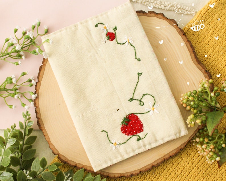 May include: A white fabric pouch with embroidered strawberries and flowers. The pouch is on a wooden slice with a yellow knitted fabric in the background.