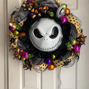 Könnte beinhalten: Ein Halloween-Kranz mit einem schwarz-weißen Jack Skellington-Gesicht, mit schwarz-weißem, orangefarbenem, gelbem, grünem und violettem Tupfenband, Mais und Spinnen-Akzenten.