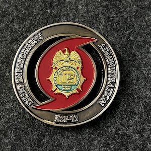 May include: A silver coin with a red and black design. The coin features a stylized hurricane with the words "Drug Enforcement Administration" and "ESF-13" around the edge. The center of the coin features a badge with the words "U.S. Drug Enforcement Administration" and "Special Agent".