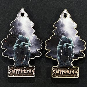 May include: Two metal air fresheners with a tree-shaped outline. Each features a dark, moody scene with a statue and the word "SUFFERING" in a decorative font. The background shows a stormy sky.