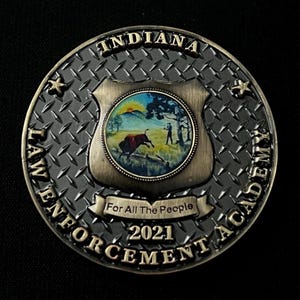May include: A round, antique-style coin with the text "INDIANA LAW ENFORCEMENT ACADEMY" and "2021". The center features a shield with a painted scene of a bull and a person. The coin has a diamond plate pattern.