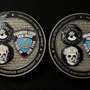 May include: Two round coins with a black border and a brick-like background. The coins feature text that reads "Special Investigations Unit" and "Organized Crime Task Force". A skull wearing a baseball cap is also visible.
