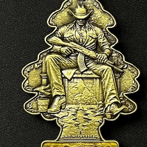 May include: A gold-colored metal air freshener with a detailed relief of a man in a cowboy hat holding a rifle. The word "NARCO" is displayed at the bottom, with "NO WITNESSES" above. The design is shaped like a stylized Christmas tree.