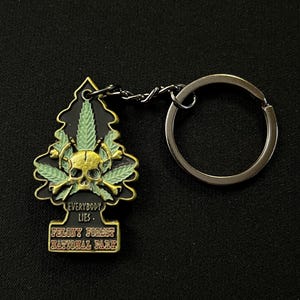 May include: A black and gold keychain with a tree-shaped design. The keychain features a skull, crossbones, and cannabis leaves. Text on the keychain reads "EVERYBODY LIES" and "FELONY FOREST NATIONAL PARK."