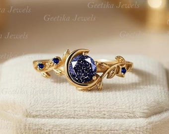 Könnte beinhalten: Ein goldfarbener Ring mit einem dunkelblauen, gesprenkelten Edelstein, der in einem Halbmond-Design gefasst ist. Der Ring hat Blatt- und kleine blaue Steinakzente am Band. Der Ring wird auf einer weißen Oberfläche präsentiert.