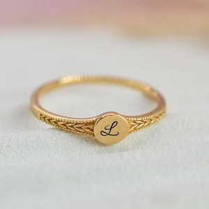 Può includere: Un anello color oro con una fascia incisa e un centro circolare. Il centro presenta la lettera "L" in corsivo nero. L'anello è posto su uno sfondo morbido e sfocato.