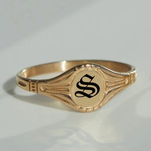 Puede incluir: Un anillo de sello de oro con una letra "S" negra grabada en una fuente inglesa antigua.