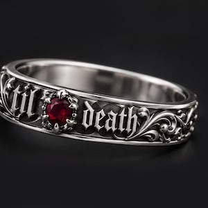 May include: A silver-toned metal ring featuring an intricate scrollwork pattern and the engraved text 'til death' in a gothic font. A single, round, deep red gemstone is set prominently between the words, accented by a dark, oxidized finish.