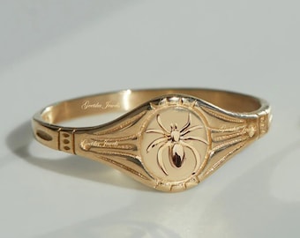 May include: A gold-colored ring with a raised spider design in the center. The ring has an oval face with decorative detailing and the text "Geetika Jewels". The ring is likely made of gold or a gold-toned metal.