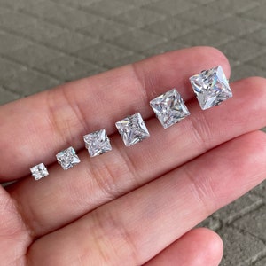 May include: A set of seven square-shaped clear cubic zirconia earrings in different sizes, displayed on a hand.