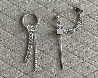 Op de afbeelding: Een paar zilveren oorbellen met ketting- en hangerontwerpen. Een oorbel heeft een hoepel met meerdere kettingen en een kleine kubusvormige hanger. De andere oorbel heeft een enkele ketting met een lange, dunne staafhanger.