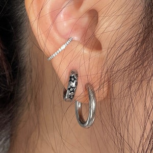 May include: A person's ear with three silver earrings. The top earring is a thin bar with small clear stones. The bottom two earrings are hoops with a star design.