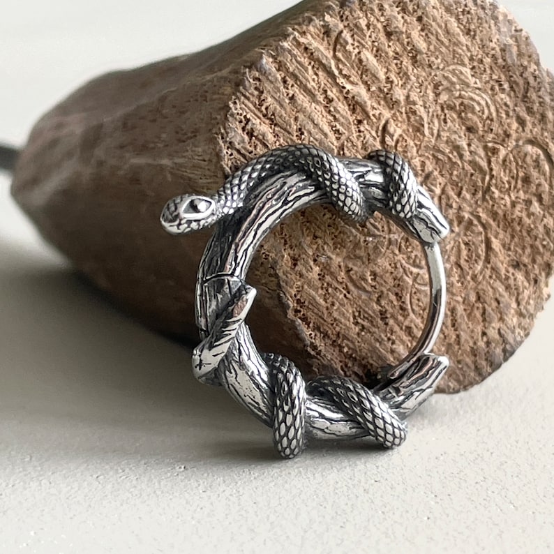 May include: A silver hoop earring with a snake coiled around a branch. The snake and branch are intricately detailed. The earring is set against a textured wooden background, showcasing the design's craftsmanship.