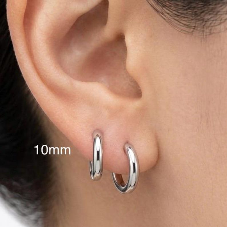 Puede incluir: Primer plano de una oreja con dos pendientes de aro plateados. Los pendientes miden 10 mm y tienen un acabado pulido. Los pendientes son de dise&ntilde;o sencillo y cl&aacute;sico.