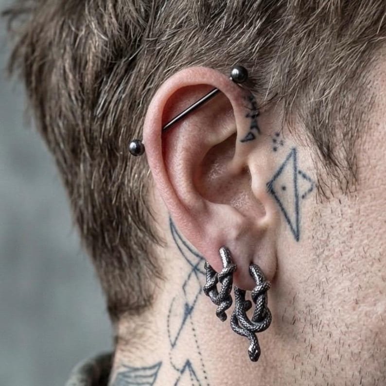 May include: Close-up of an ear featuring a silver industrial barbell piercing and two pairs of silver snake hoop earrings. The ear is pierced and surrounded by tattoos on the skin.