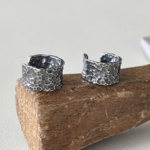 May include: Two silver ear cuffs with a hammered texture. The cuffs have an open design and a dark, oxidized finish. They are displayed on a piece of wood against a neutral background. The jewellery showcases the texture and detail of the metal.
